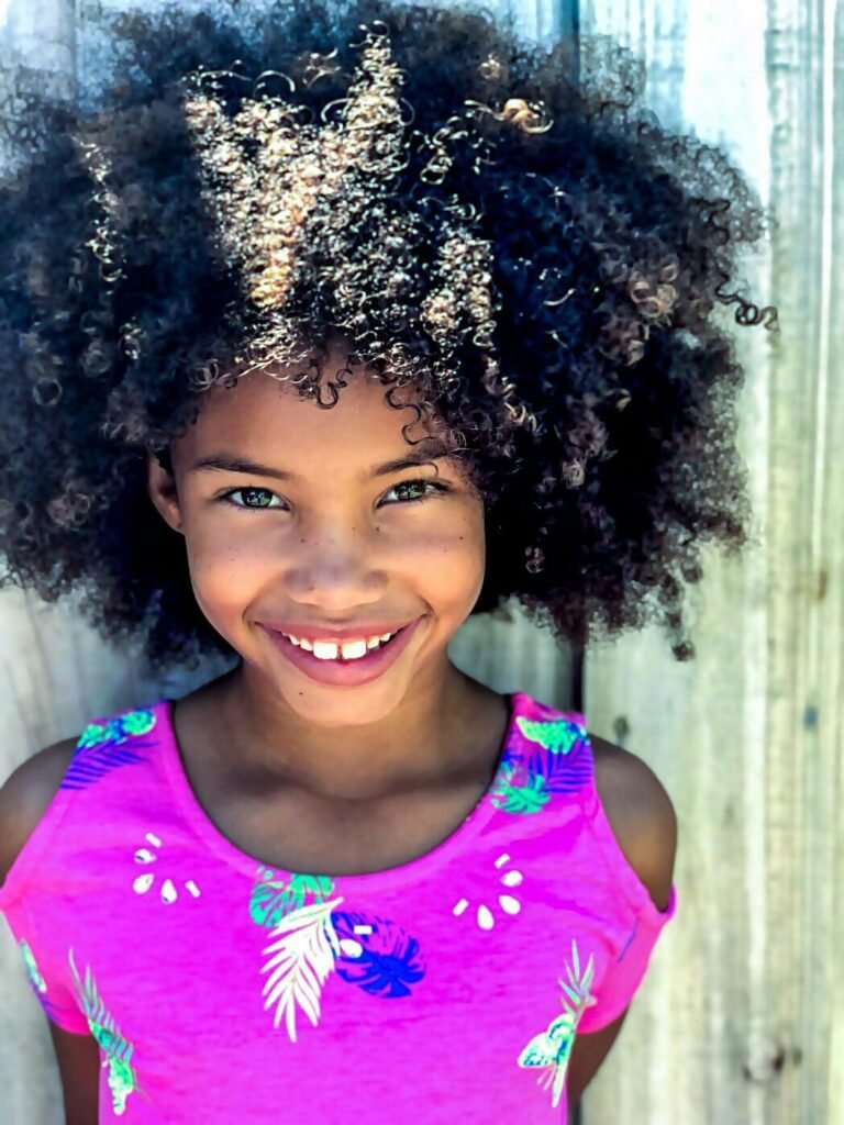 pexels photo 1068205 1068205 A joyful young girl showcases her beautiful curly Afro hair and bright smile in an outdoor portrait against a wooden backdrop.